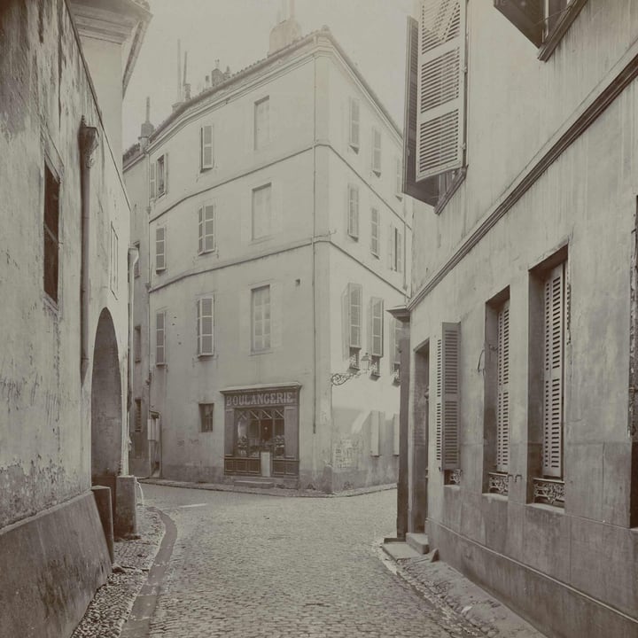 Before: a scanned 1908 print of a narrow cobbled street with a bakery on the corner, flat and brownish, with a grainy surface