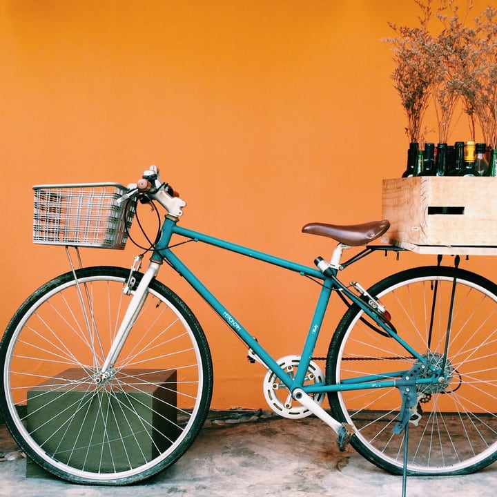 Before: a still photo of a teal bicycle with a basket against an orange wall, with a crate of bottles and dried flowers behind it